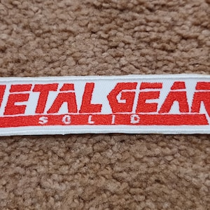 May include: A rectangular embroidered patch featuring the red text METAL GEAR on a white background, with the word SOLID in smaller red letters centered below. The patch has a white border and is designed for attachment to fabric items.