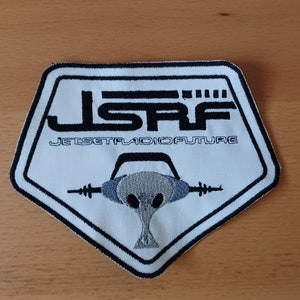 May include: A pentagonal embroidered patch featuring a white background with a black border. The design includes the black text JSRF and JET SET RADIO FUTURE, along with a central grey and black graphic of a stylized character wearing headphones.