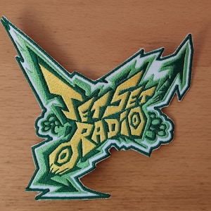 May include: An embroidered patch featuring the text JET SET RADIO in yellow block letters. The design is surrounded by jagged, lightning-like shapes in various shades of green and white, creating a dynamic, angular, and stylized graphic emblem.