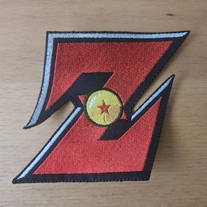 May include: An embroidered patch in the shape of a stylized letter Z, featuring a vibrant red center with black borders and a thin light blue outline. Centered on the patch is a circular yellow sphere with a single red star and a white highlight.