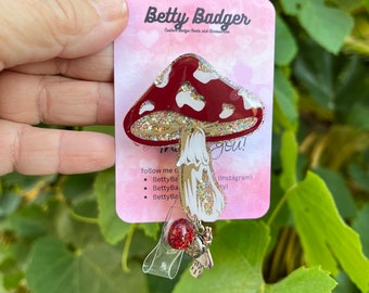 May include: A red and white mushroom-shaped badge reel with glitter accents. The badge reel has a clear plastic retractable cord and a charm with a red glitter ball and a small bee. The card reads "Betty Badger" and "follow me".
