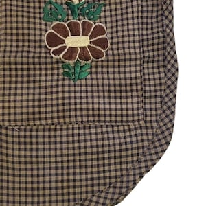 Puede incluir: Primer plano de un delantal a cuadros marr&oacute;n y beige con un dise&ntilde;o de bordado floral. La flor bordada es marr&oacute;n con un centro crema y hojas verdes. El delantal tiene un bolsillo y un borde inferior curvo.