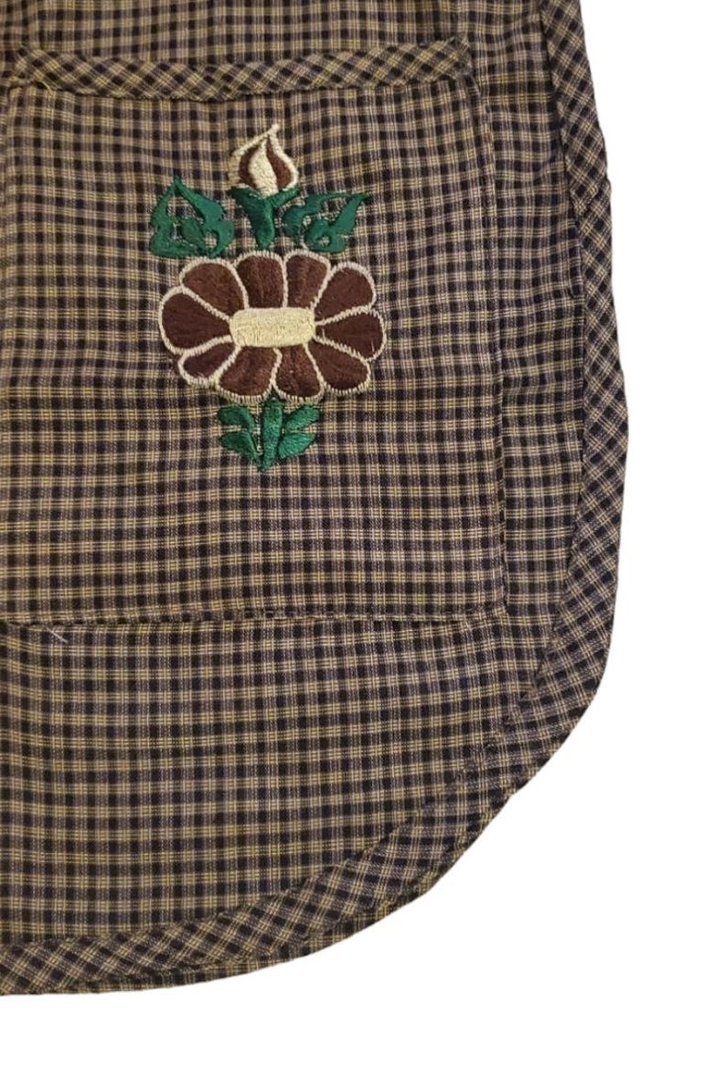 Puede incluir: Primer plano de un delantal a cuadros marr&oacute;n y beige con un dise&ntilde;o de bordado floral. La flor bordada es marr&oacute;n con un centro crema y hojas verdes. El delantal tiene un bolsillo y un borde inferior curvo.