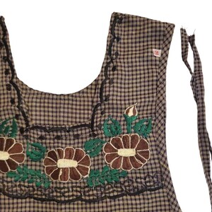 Puede incluir: Un delantal a cuadros marrones y negros con detalles florales bordados. El delantal presenta flores marrones con centros crema y hojas verdes. Las costuras decorativas negras bordean el escote y los bordes. El delantal tiene correas.