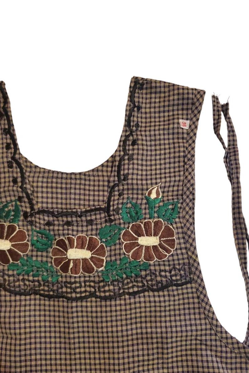 Puede incluir: Un delantal a cuadros marrones y negros con detalles florales bordados. El delantal presenta flores marrones con centros crema y hojas verdes. Las costuras decorativas negras bordean el escote y los bordes. El delantal tiene correas.