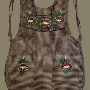 May include: A checkered apron with embroidered floral designs. The apron has a brown and white checkered pattern, with brown and green embroidered flowers on the chest and pockets. It has ties at the neck and waist.