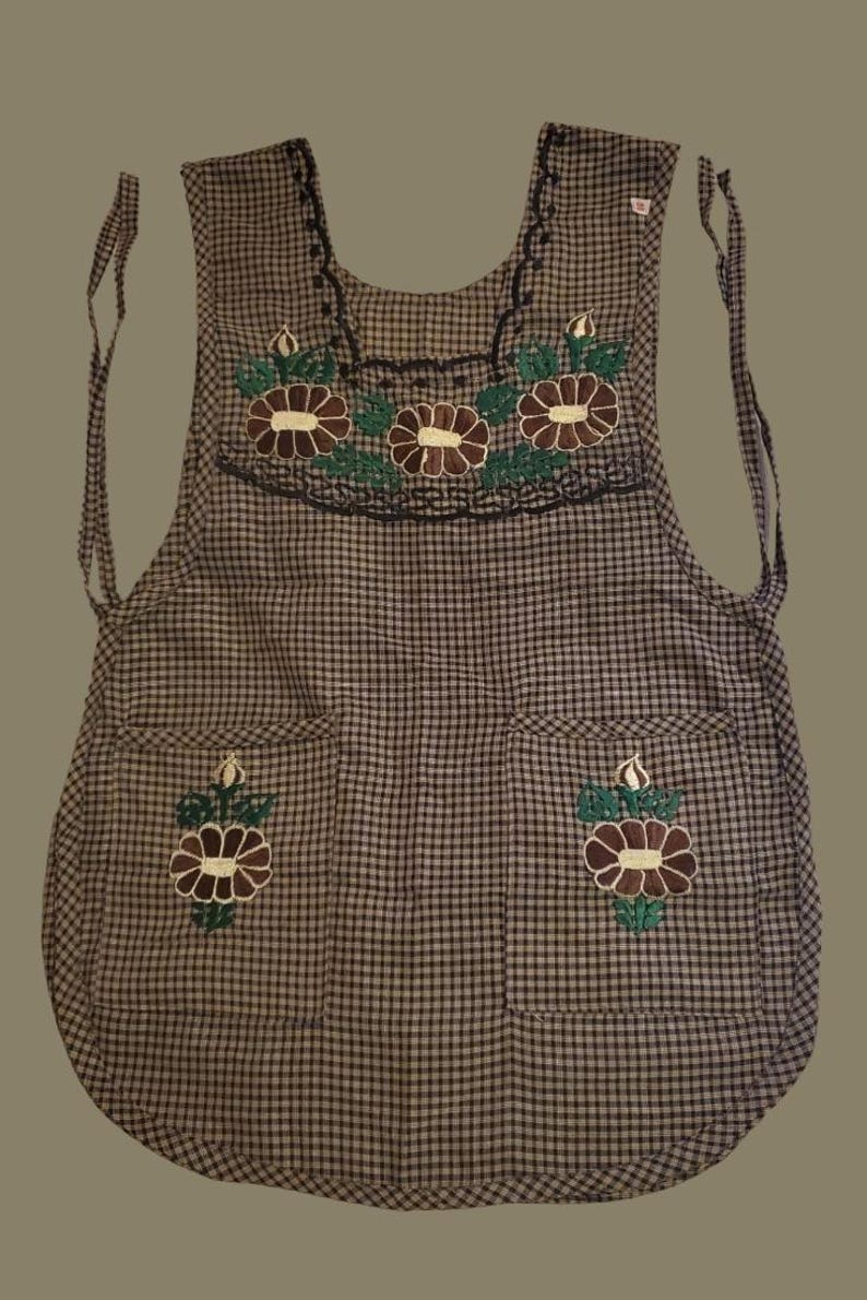 Puede incluir: Un delantal a cuadros con dise&ntilde;os florales bordados. El delantal presenta un patr&oacute;n a cuadros marr&oacute;n y blanco, con flores bordadas marrones y verdes en el pecho y los bolsillos. Tiene lazos en el cuello y la cintura.