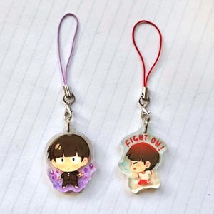 May include: Two anime character charms with purple and red strings. The charm on the left is a boy with black hair and a purple background. The charm on the right is a boy with red hair and a white background with the text "FIGHT ON!" in red.