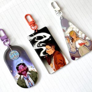 May include: Three colourful keychains with different designs. The first keychain features a disco ball and a man with a halo. The second keychain features a man with a cigarette and a black and white smoke design. The third keychain features a woman with a red heart and a white dress.
