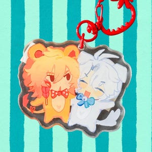 May include: A clear acrylic charm featuring two cartoon characters. One is a yellow bear-like figure holding a lollipop, and the other is a white dog-like character. Both are smiling and wearing bow ties. The charm has a red clasp.