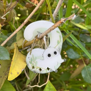 May include: A white enamel pin in the shape of a stylized elephant with a curved trunk. The pin features black eyes, small feet, and green leaf accents. It is hanging on a small branch, surrounded by green foliage.