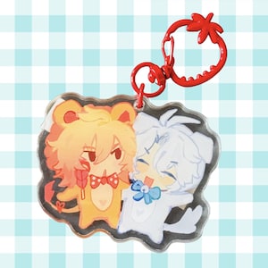 May include: An acrylic keychain featuring two cartoon characters: a yellow lion-like figure with a red polka-dot bowtie and a white dog-like figure with a blue bowtie. The keychain has a red clasp shaped like a strawberry at the top.