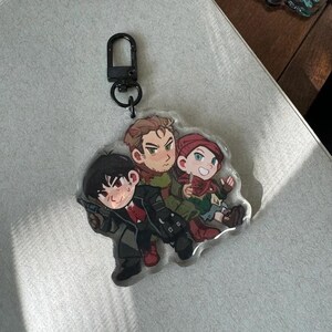 May include: A clear acrylic keychain featuring three cartoon characters. One character is holding a gun, another is wearing a red beanie, and the third is wearing a green scarf. The keychain has a black clasp.