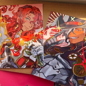 May include: Two art prints with detailed character illustrations. One shows a character with red hair and roses, the other a character in a military uniform with a hat. Both prints have a vibrant colour palette with gold accents, creating a striking visual.