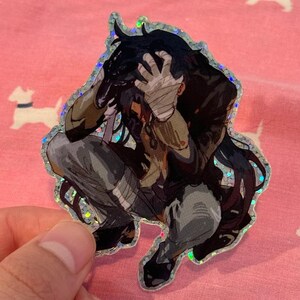 May include: A holographic sticker featuring a black-haired anime character with a distressed expression, wearing a brown jacket and grey pants. The character is holding their head in their hands, with bandages wrapped around their hands.