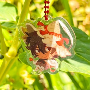 May include: A clear acrylic charm depicting a cartoon character with brown hair and a festive outfit. The charm is attached to a red beaded chain. The character has a tan and red outfit with glitter accents. The charm is hanging in front of green foliage.