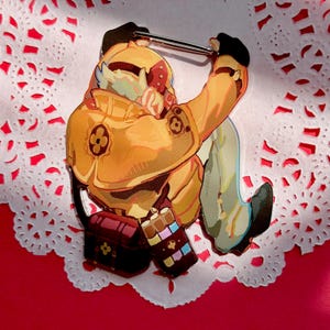 May include: A colorful pin featuring a cartoon character in a yellow jacket, black gloves, and light green pants. The character is holding a pin. A red treasure chest and a chocolate bar are also featured in the design. The pin is on a white doily.