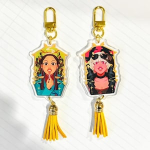 May include: Two colorful acrylic keychains with gold-colored metal rings and tassels. The keychains feature illustrations of two women with different hairstyles and clothing. One woman is wearing a blue shirt and has a lipstick in her hand. The other woman is wearing a black leather jacket and sunglasses.