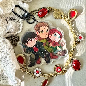 Puede incluir: Un llavero acrílico transparente con una imagen de dibujos animados de tres personajes. El llavero está unido a un cierre negro y una cadena dorada adornada con joyas rojas y adornos en forma de flor.