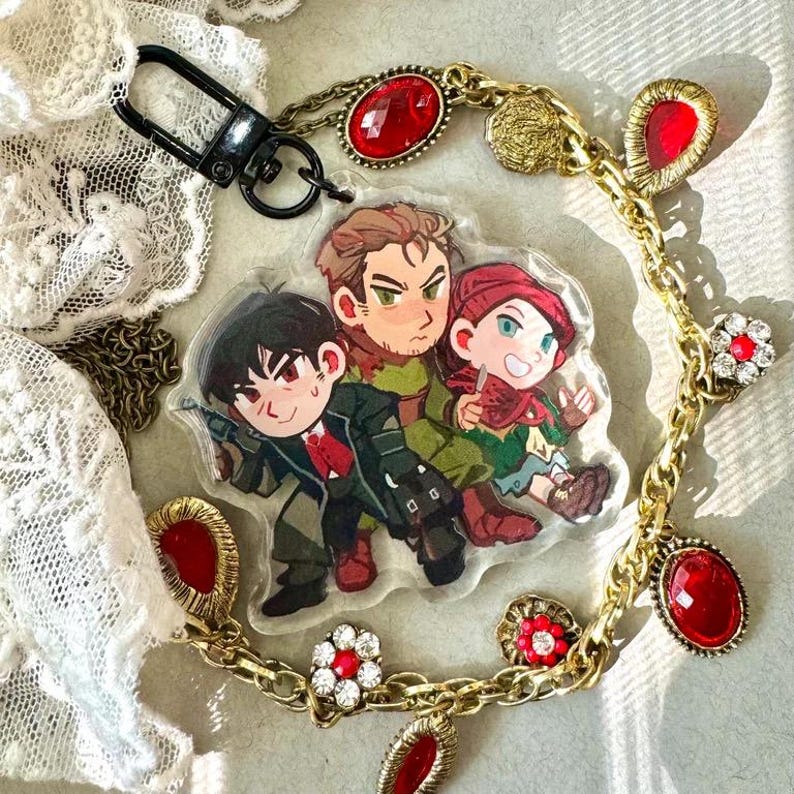 May include: A clear acrylic keychain featuring a cartoon image of three characters. The keychain is attached to a black clasp and a gold chain adorned with red jewels and flower-shaped embellishments.