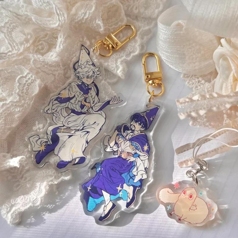 May include: Two colourful anime character keychains with gold chains and a smaller white keychain with a gold chain. The characters are a boy with a white hat and a boy with a blue hat. The smaller keychain is a white animal with a pink nose.