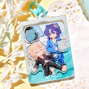 May include: A clear acrylic keyring with two anime-style characters in a sleeping scene. One character has blue hair and wears a blue robe, while the other has blonde hair and wears a black outfit. The keyring has a blue clasp.