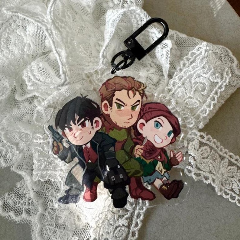 May include: A clear acrylic charm featuring three cartoon characters. One character holds a gun, another has a knife, and the third has a black bag. The charm has a black clasp attached. The background is white lace.