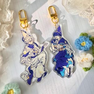 May include: Two colourful anime-style character charms with gold keychains. The charms are made of clear acrylic and feature a blue and white character with a pointed hat and a white and gold character with a pointed hat. Both characters are holding a wand.