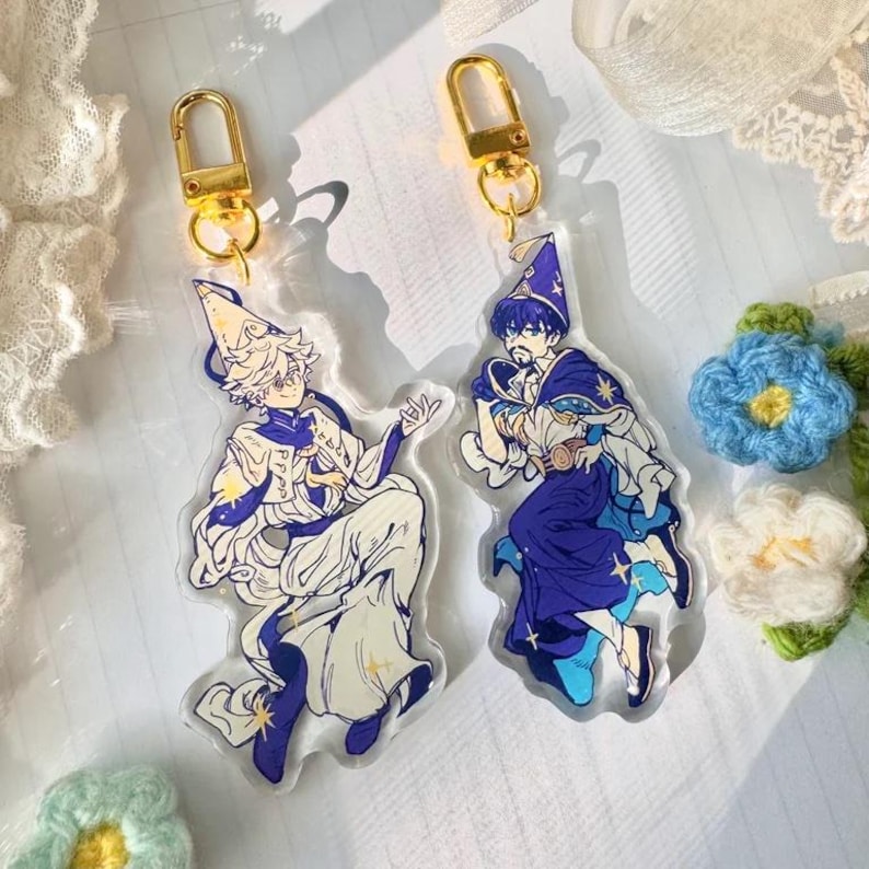 May include: Two colourful anime-style character charms with gold keychains. The charms are made of clear acrylic and feature a blue and white character with a pointed hat and a white and gold character with a pointed hat. Both characters are holding a wand.