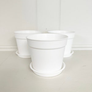 May include: Three white plastic plant pots with matching saucers. The pots are all the same size and are arranged in a row.