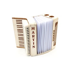 Könnte beinhalten: Ein helles Holzakkordeon mit dem vertikal angebrachten Namen "MARTIN". Das Instrument hat eine Tastatur auf der linken Seite und einen Balgabschnitt mit weißen Seiten. Es hat braune Akzente.