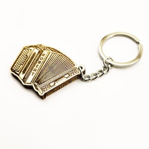 Op de afbeelding: Een houten sleutelhanger met een gedetailleerde accordeon met het woord "ROBERT" erop gegraveerd. De sleutelhanger is bevestigd aan een zilveren metalen ring en ketting.