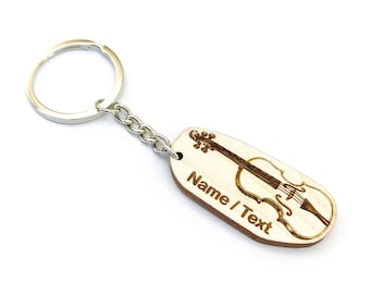 Puede incluir: Un llavero de madera con una cadena y un anillo plateados. La etiqueta de madera de color claro tiene un diseño de violín grabado con láser y el texto "Name / Text". El llavero está sobre un fondo blanco.