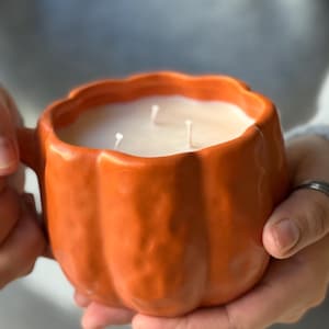 May include: An orange pumpkin-shaped candle holder filled with white wax and three wicks. The ceramic holder has a handle and is held in both hands. The candle is a fall-themed decorative item.