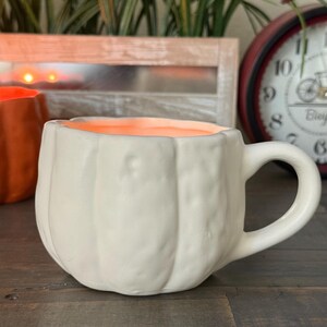 May include: A white ceramic mug shaped like a pumpkin with a handle. The interior of the mug is orange. The mug is sitting on a wooden surface. A second orange pumpkin-shaped mug is partially visible in the background.