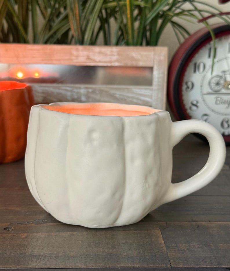 May include: A white ceramic mug shaped like a pumpkin with a handle. The interior of the mug is orange. The mug is sitting on a wooden surface. A second orange pumpkin-shaped mug is partially visible in the background.