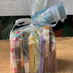 May include: A clear plastic bag filled with a variety of handmade soap bars. The bars are arranged in a stack and are wrapped with a silver ribbon. A gift tag with the text "Merry Christmas" is attached to the bag.