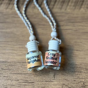 Puede incluir: Dos peque&ntilde;os frascos de vidrio con tapas blancas y cordones colgantes. Un frasco tiene una etiqueta que dice "Sugared Pumpkin" y el otro "Cozy Knitted Sweater". Los frascos est&aacute;n sobre una superficie de madera.