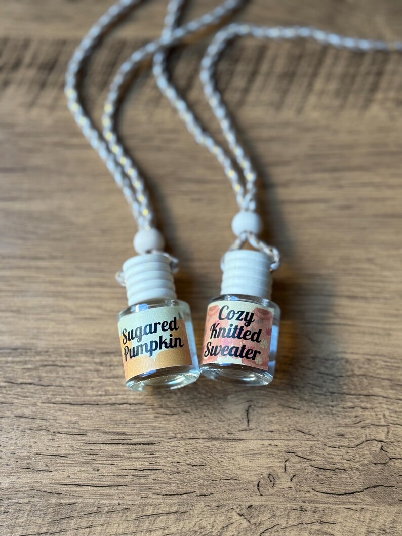 Puede incluir: Dos peque&ntilde;os frascos de vidrio con tapas blancas y cordones colgantes. Un frasco tiene una etiqueta que dice "Sugared Pumpkin" y el otro "Cozy Knitted Sweater". Los frascos est&aacute;n sobre una superficie de madera.