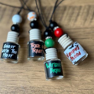 May include: Four small, clear glass bottles with black labels and white text, each with a wooden top and a beaded string. The labels read "Ghost With The Most", "Trick or Treats", "Witches Brew", and "Vampire Blood". The beads are black, white, orange, red, and green.