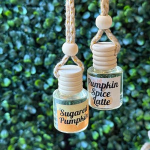 Puede incluir: Dos peque&ntilde;as botellas de vidrio con tapas de madera y cordones colgantes. Una botella tiene una etiqueta que dice "Sugared Pumpkin" y la otra "Pumpkin Spice Latte". Las botellas cuelgan frente a un fondo verde frondoso.
