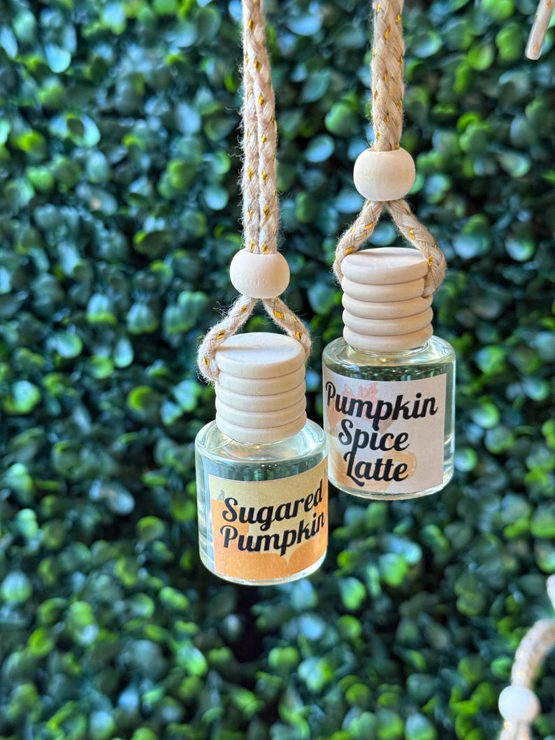 Puede incluir: Dos peque&ntilde;as botellas de vidrio con tapas de madera y cordones colgantes. Una botella tiene una etiqueta que dice "Sugared Pumpkin" y la otra "Pumpkin Spice Latte". Las botellas cuelgan frente a un fondo verde frondoso.