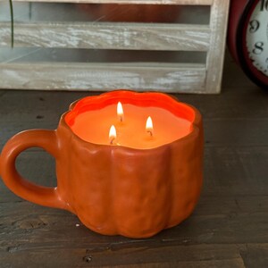 May include: An orange, pumpkin-shaped candle with three lit wicks. The candle is in a mug with a handle, sitting on a wooden surface. The background includes a wooden crate with greenery and a red clock.