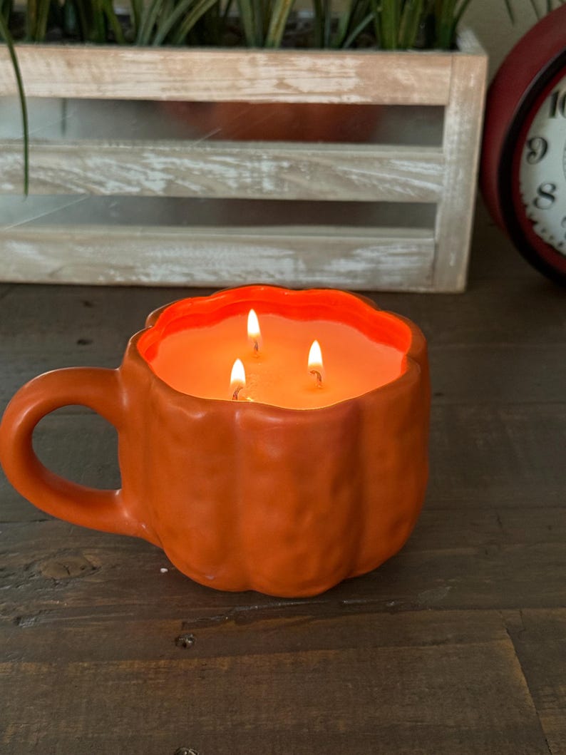 May include: An orange, pumpkin-shaped candle with three lit wicks. The candle is in a mug with a handle, sitting on a wooden surface. The background includes a wooden crate with greenery and a red clock.