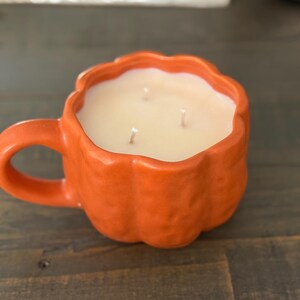 May include: An orange ceramic candle shaped like a pumpkin mug. The mug has a handle and is filled with a light-colored wax. Three wicks are visible. The candle sits on a dark wooden surface.