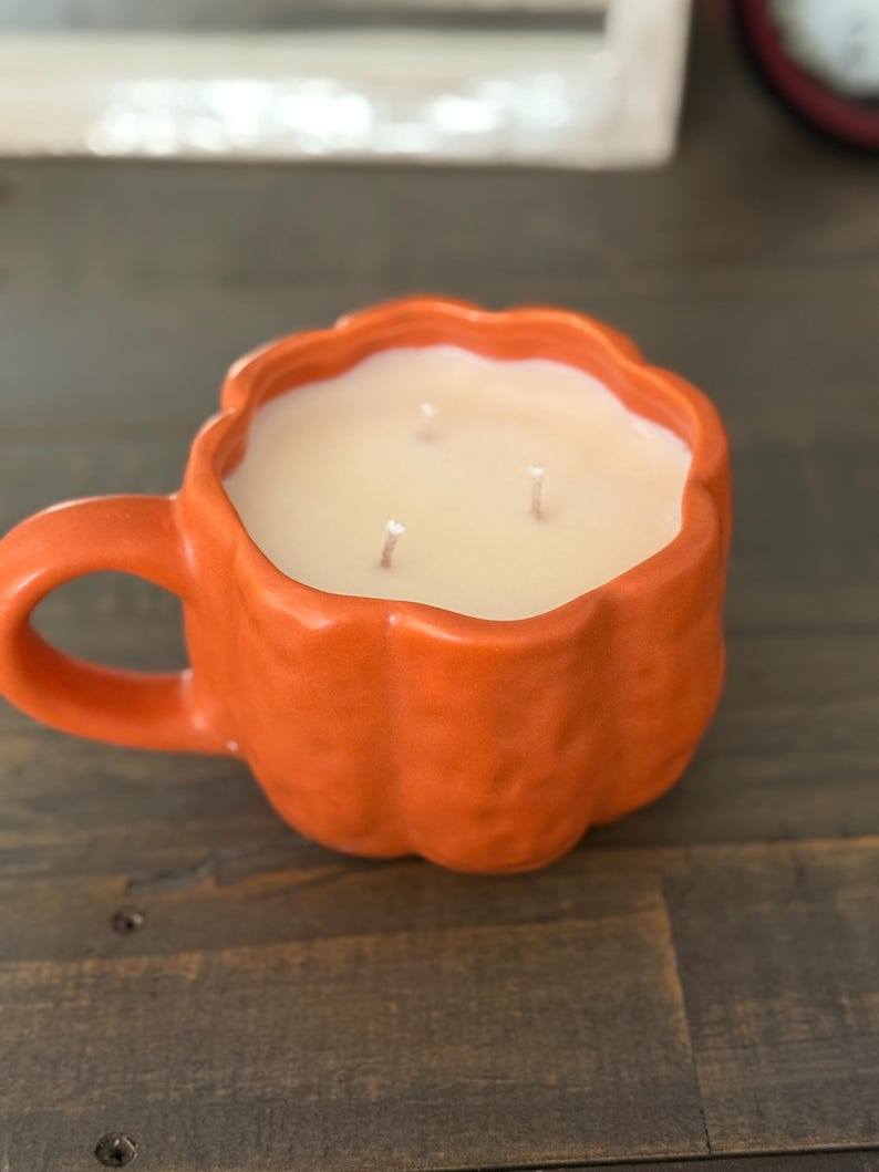 May include: An orange ceramic candle shaped like a pumpkin mug. The mug has a handle and is filled with a light-colored wax. Three wicks are visible. The candle sits on a dark wooden surface.