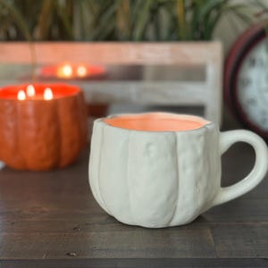 May include: A white ceramic mug shaped like a pumpkin with a handle, sitting on a wooden surface. The interior of the mug is orange. In the background, another orange pumpkin-shaped candle is visible, along with a red clock.