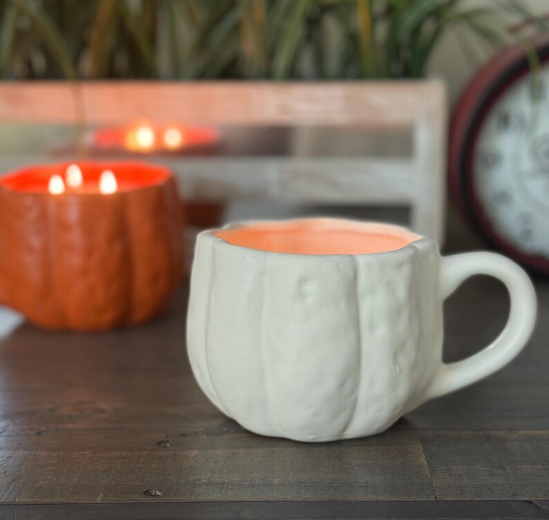 May include: A white ceramic mug shaped like a pumpkin with a handle, sitting on a wooden surface. The interior of the mug is orange. In the background, another orange pumpkin-shaped candle is visible, along with a red clock.