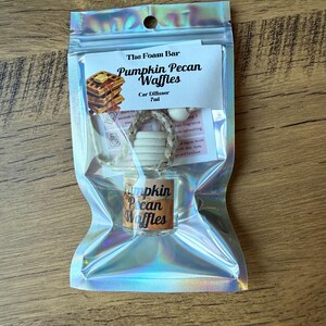 Puede incluir: Un ambientador para coche en una bolsa hologr&aacute;fica. La etiqueta dice "Pumpkin Pecan Waffles" con una imagen de gofres. El producto incluye un peque&ntilde;o frasco de vidrio y un difusor de madera. La bolsa est&aacute; sellada y lista para usar.