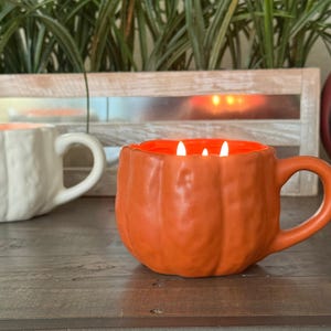 May include: An orange pumpkin-shaped mug candle with three lit wicks. The interior of the mug is orange, and the exterior has a textured pumpkin design. A white pumpkin-shaped mug is in the background. The mugs are on a wooden surface.
